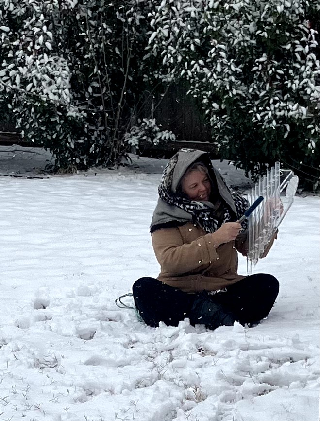 Child sitting in snow, holding a plastic container, with snow-covered trees in background.