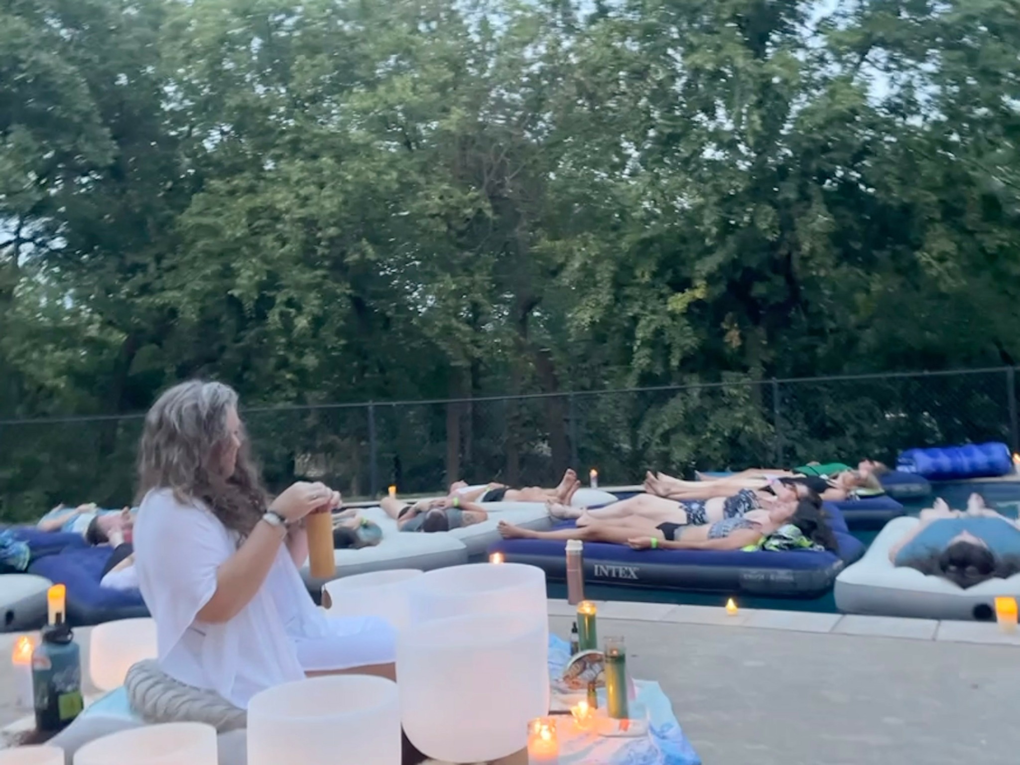 Group of people lying on inflatable mattresses outdoors, surrounded by trees, with some standing and drinking.