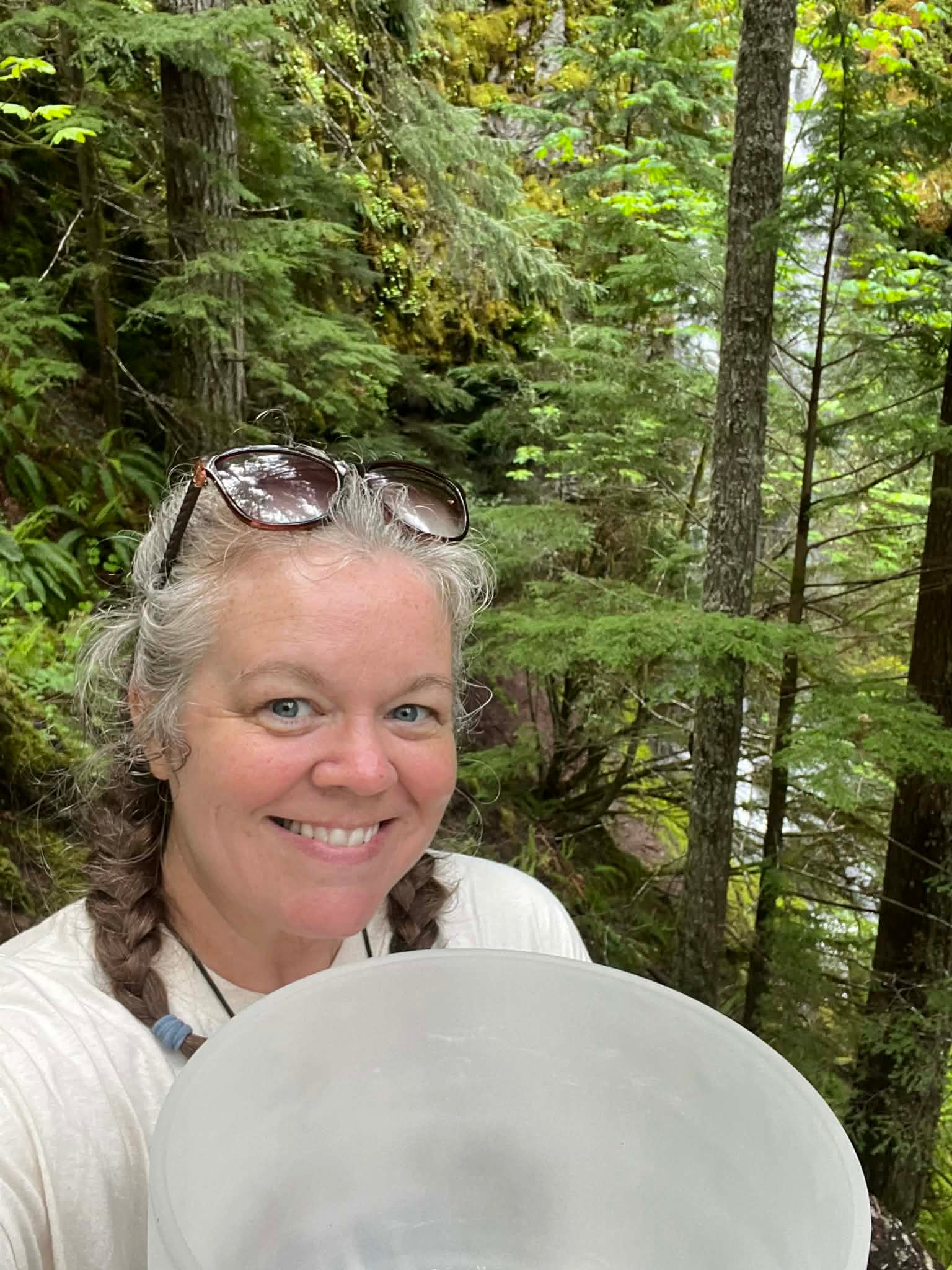 Smiling woman with braided hair and sunglasses on head in a forest with green trees.