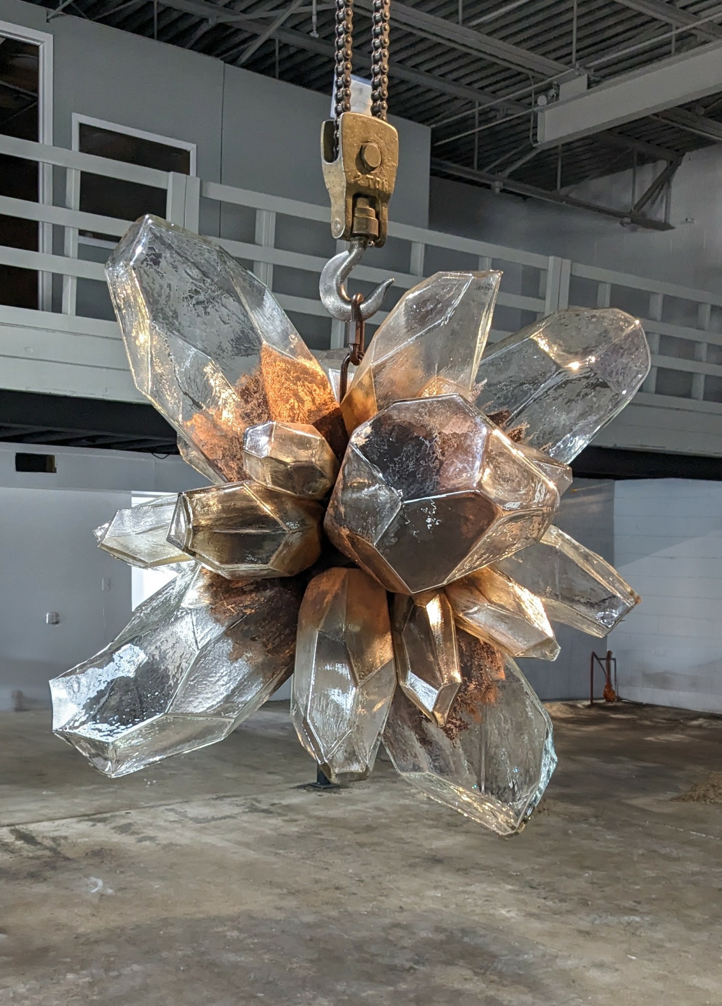 Large crystal-like sculpture hanging from a chain in an industrial space with concrete floor and metal ceiling.