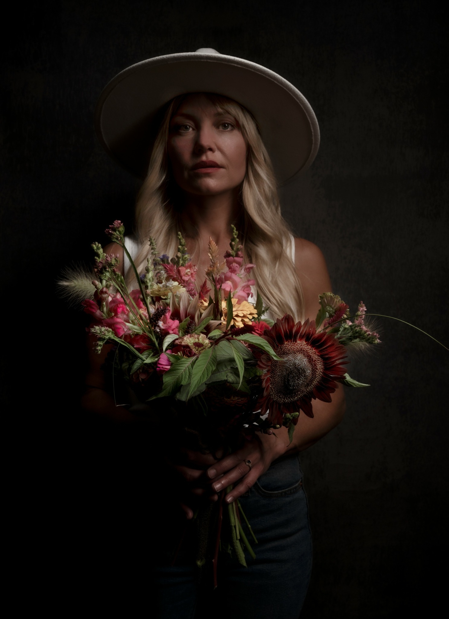 Woman with long blonde hair wearing a wide-brimmed hat holding a large colorful flower bouquet.