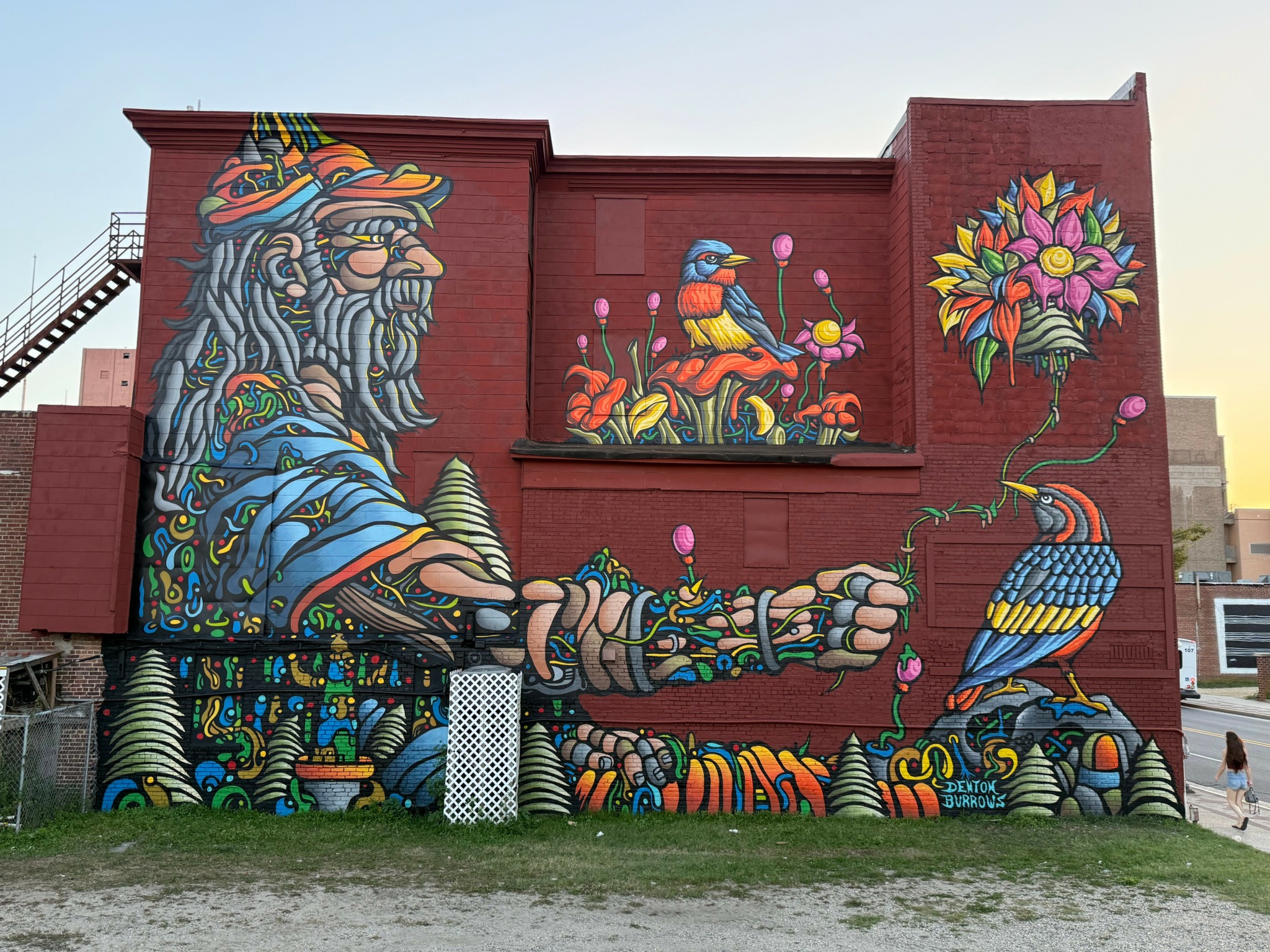 Colorful mural of a bearded figure with a long arm holding a bird, surrounded by flowers and vibrant patterns on a red brick wall.
