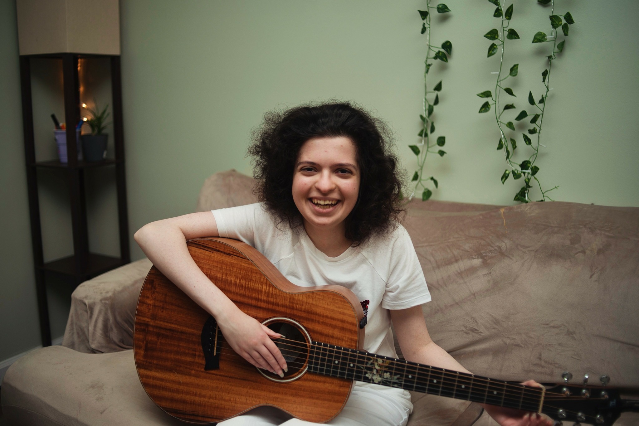 Woman with curly hair smiling, holding an acoustic guitar on a sofa in a room with green wall and plants.