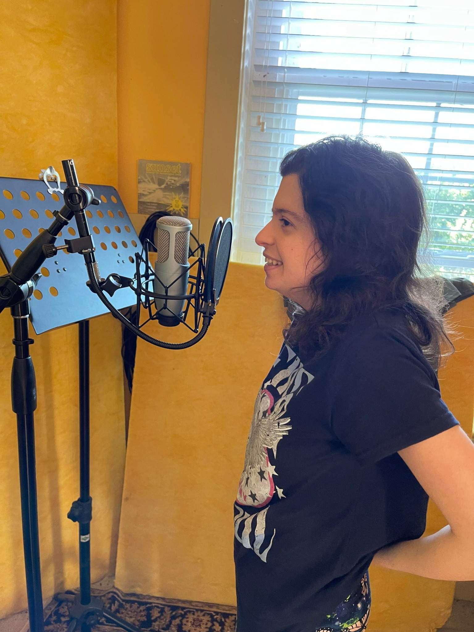 Girl with dark hair in a black shirt, standing indoors near a window with curtains, microphone stand nearby.