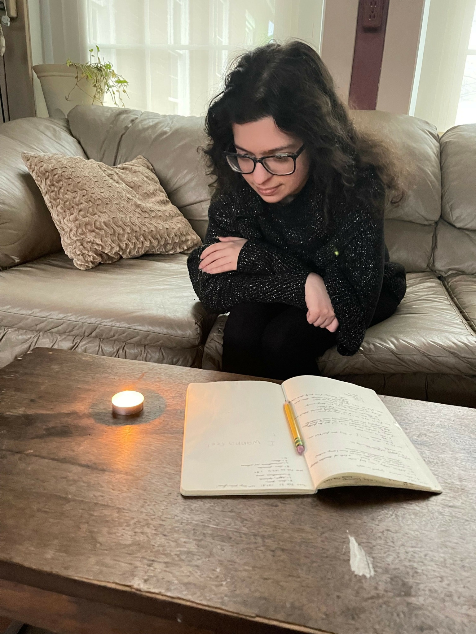 Woman with glasses and dark hair reading a book on a couch with a candle on the table.