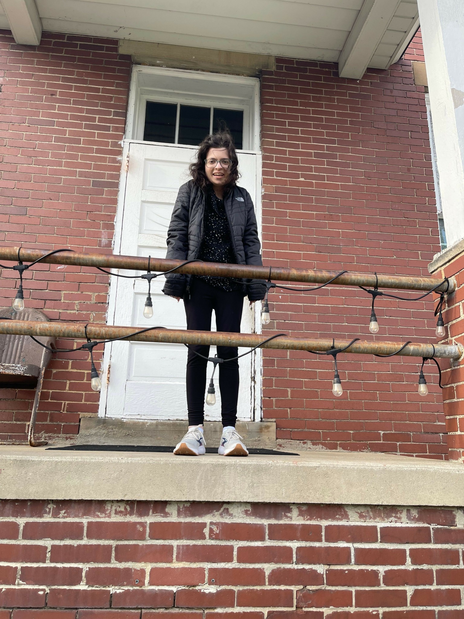 Person standing outside brick building near door, wearing dark jacket and pants, with a small tree in front.