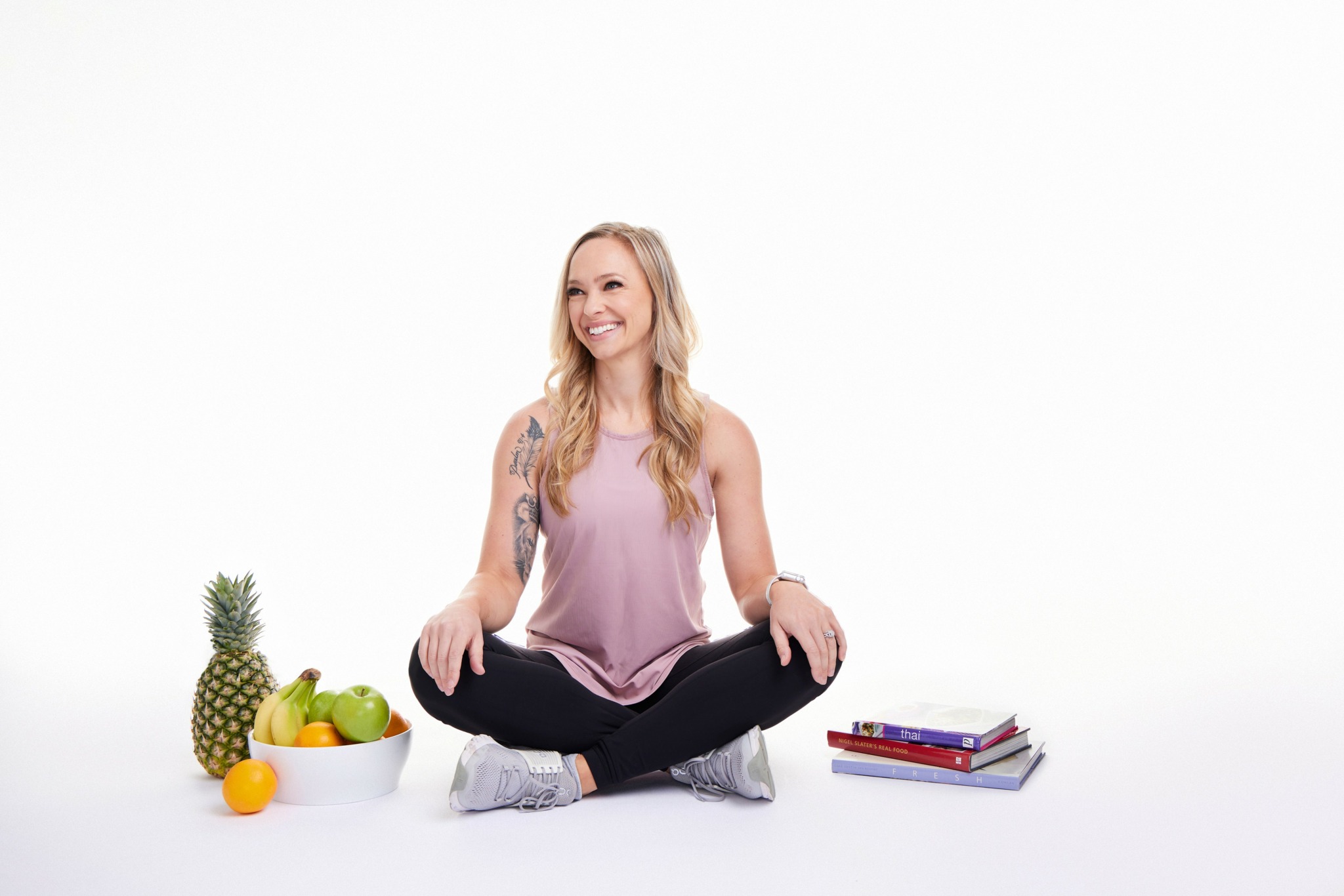 Woman sitting cross-legged with a smile, surrounded by a pineapple, fruit bowl, and books, on a white background.