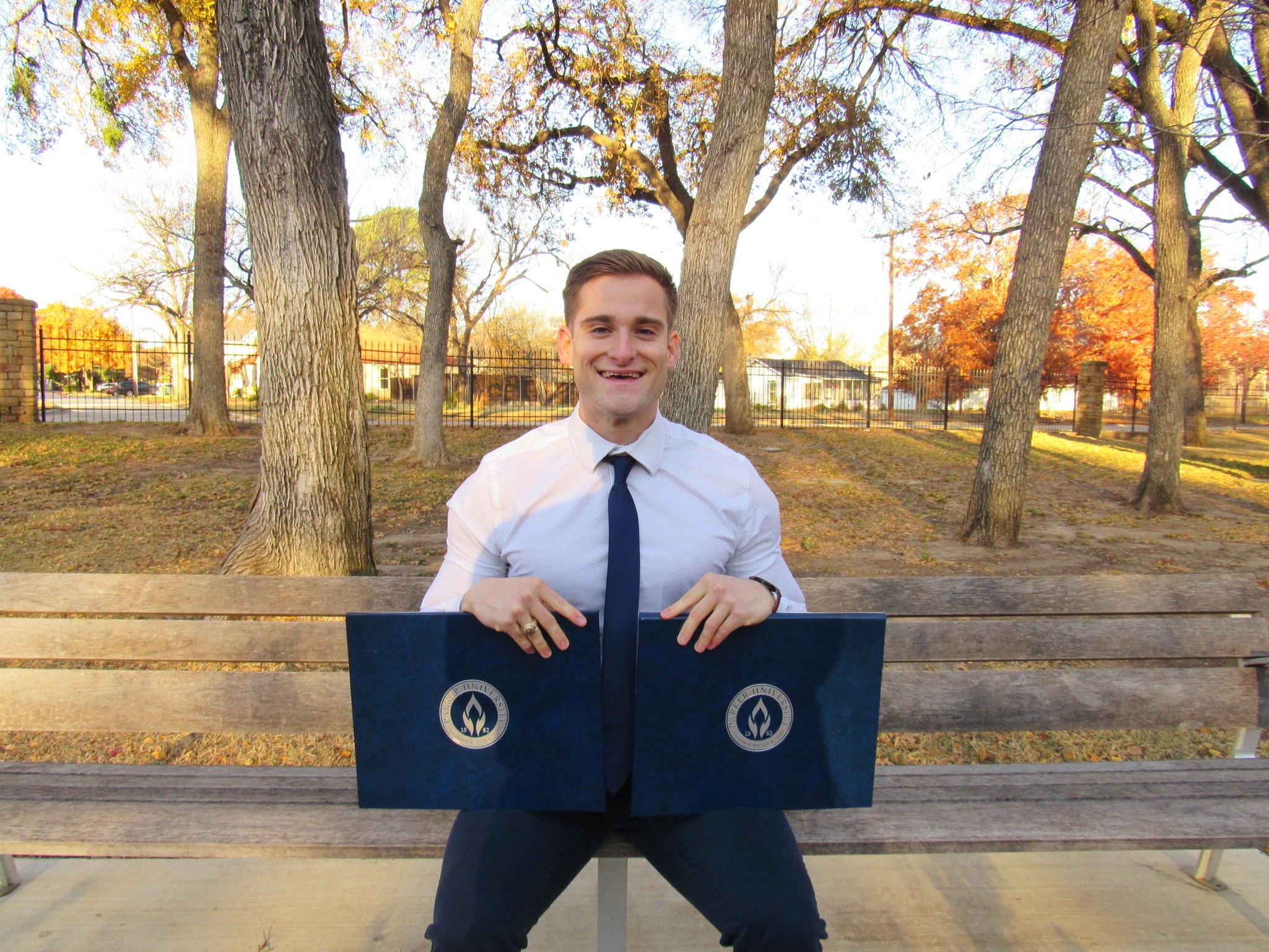 Young man in formal attire sitting on a park bench holding an open folder outdoors with trees and autumn foliage.