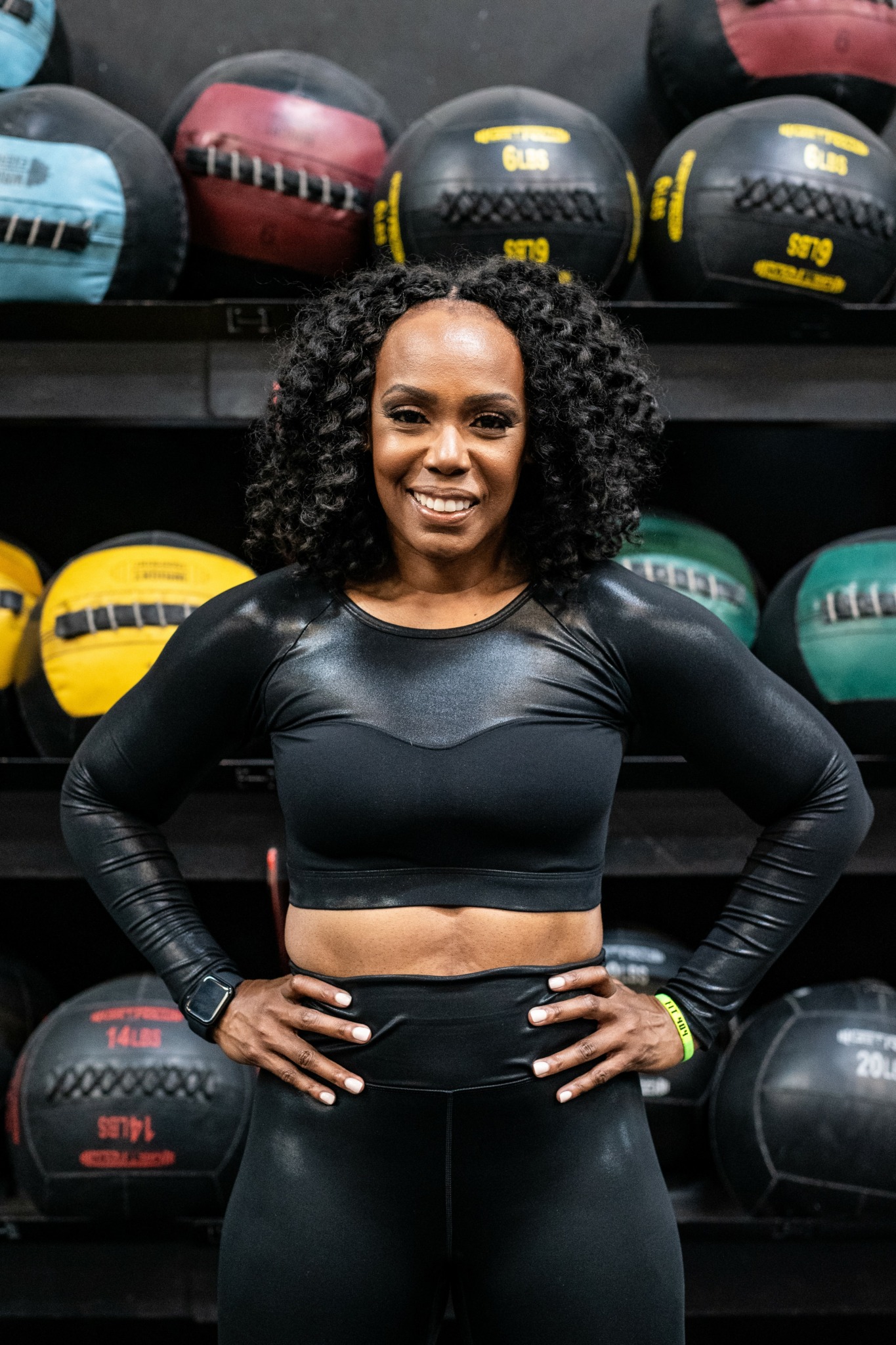 Woman with curly hair in black workout clothes smiling in gym with medicine balls behind her.