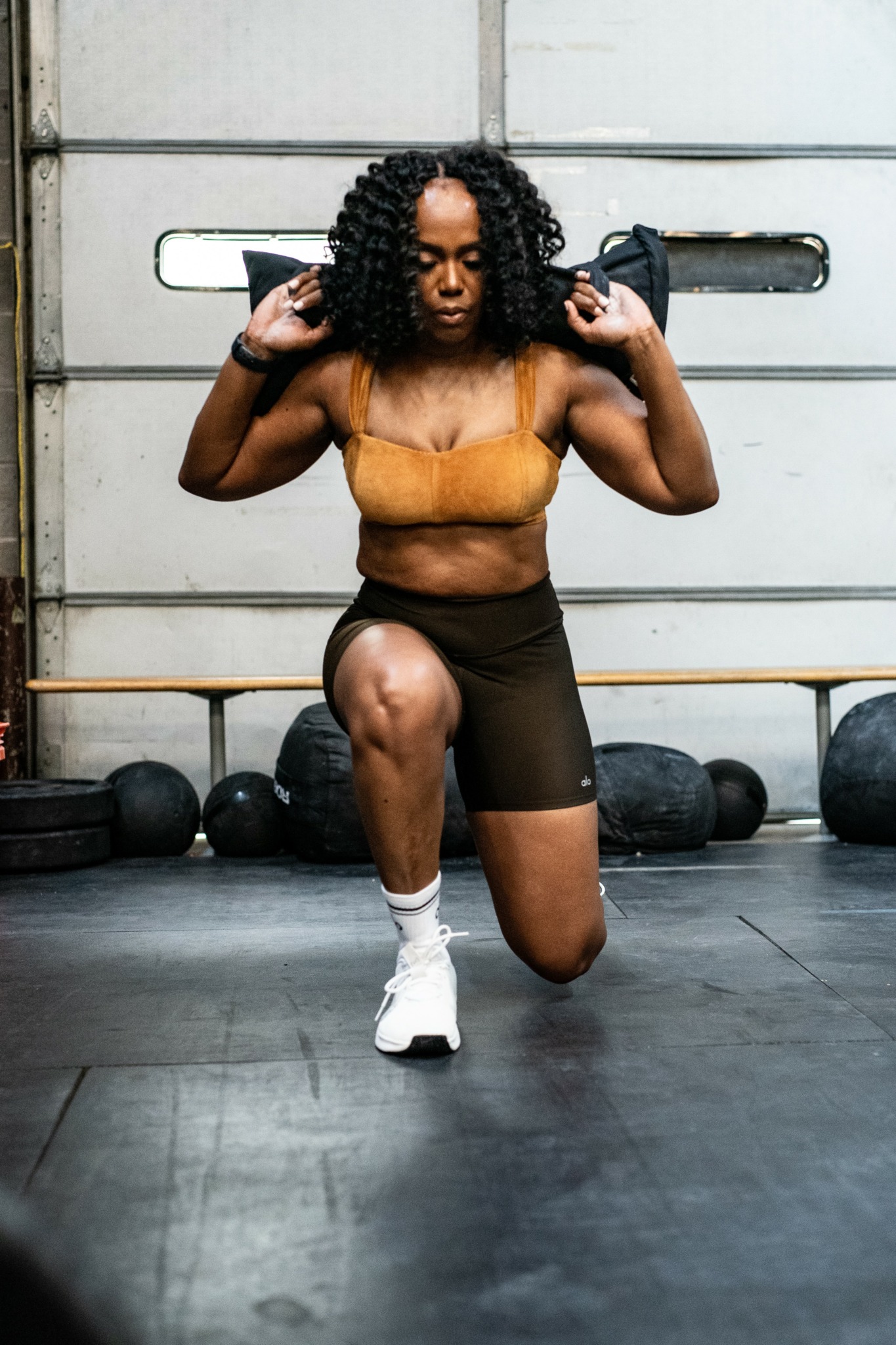 Woman with curly hair in workout clothes kneeling in gym, holding weights on shoulders.