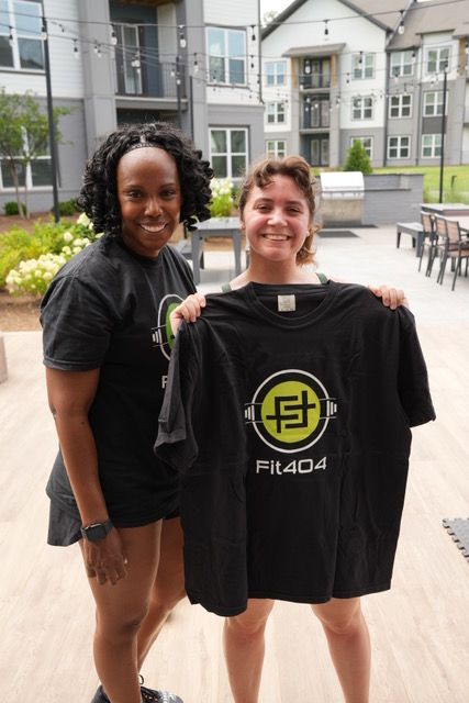Two women standing outdoors in front of modern buildings, smiling, one holding a black bag with a logo, casual clothing.