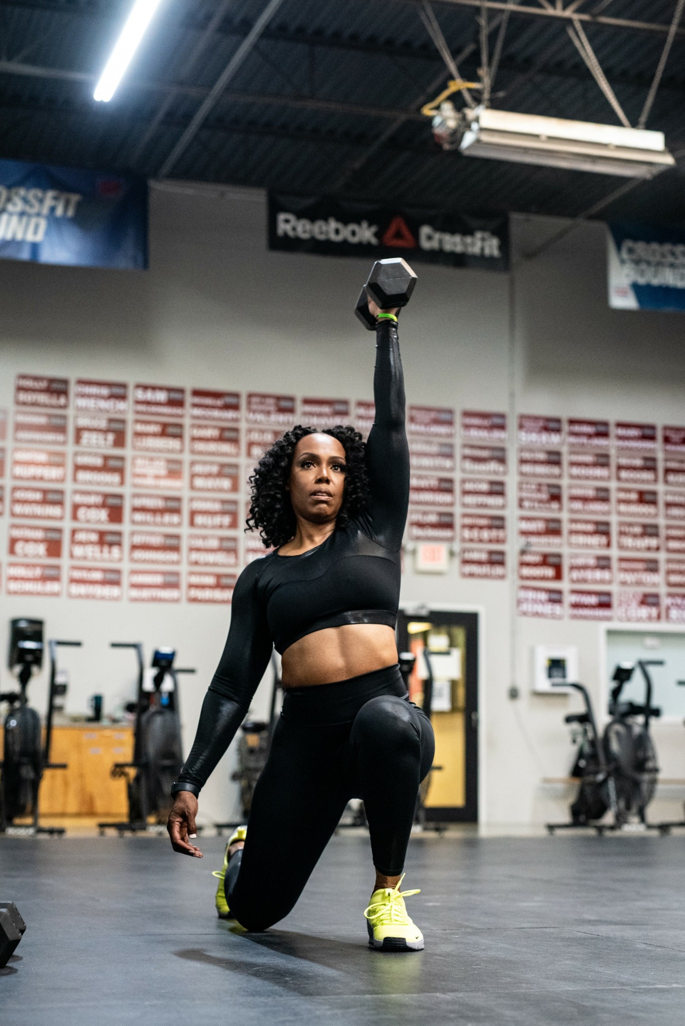 Woman in black workout clothes kneels on one knee in a gym, holding a kettlebell overhead with one arm.