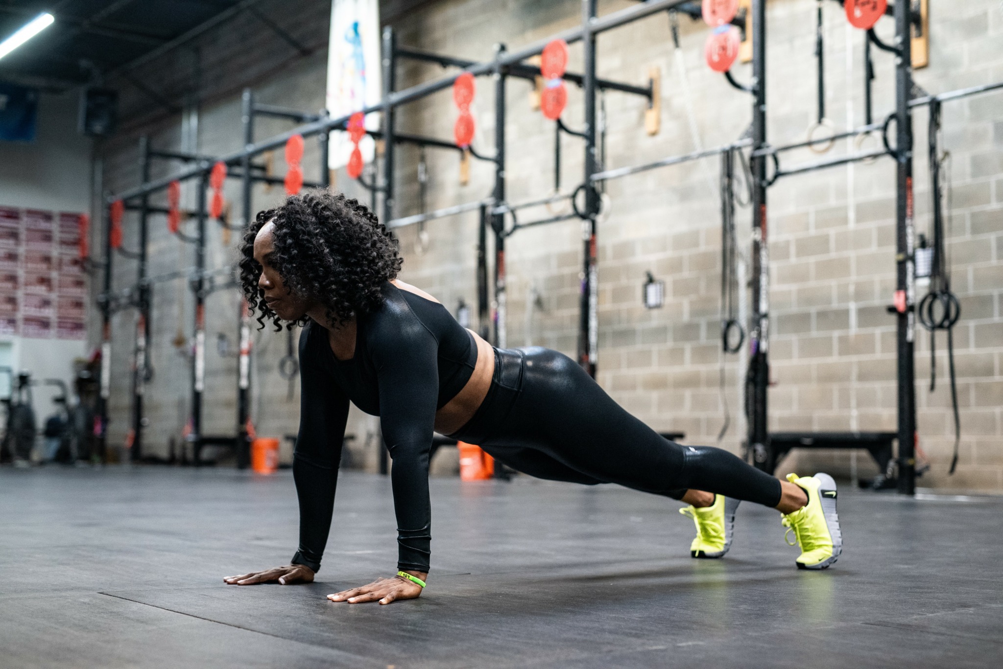 Woman performing a plank exercise in a gym with workout equipment and brick wall background.