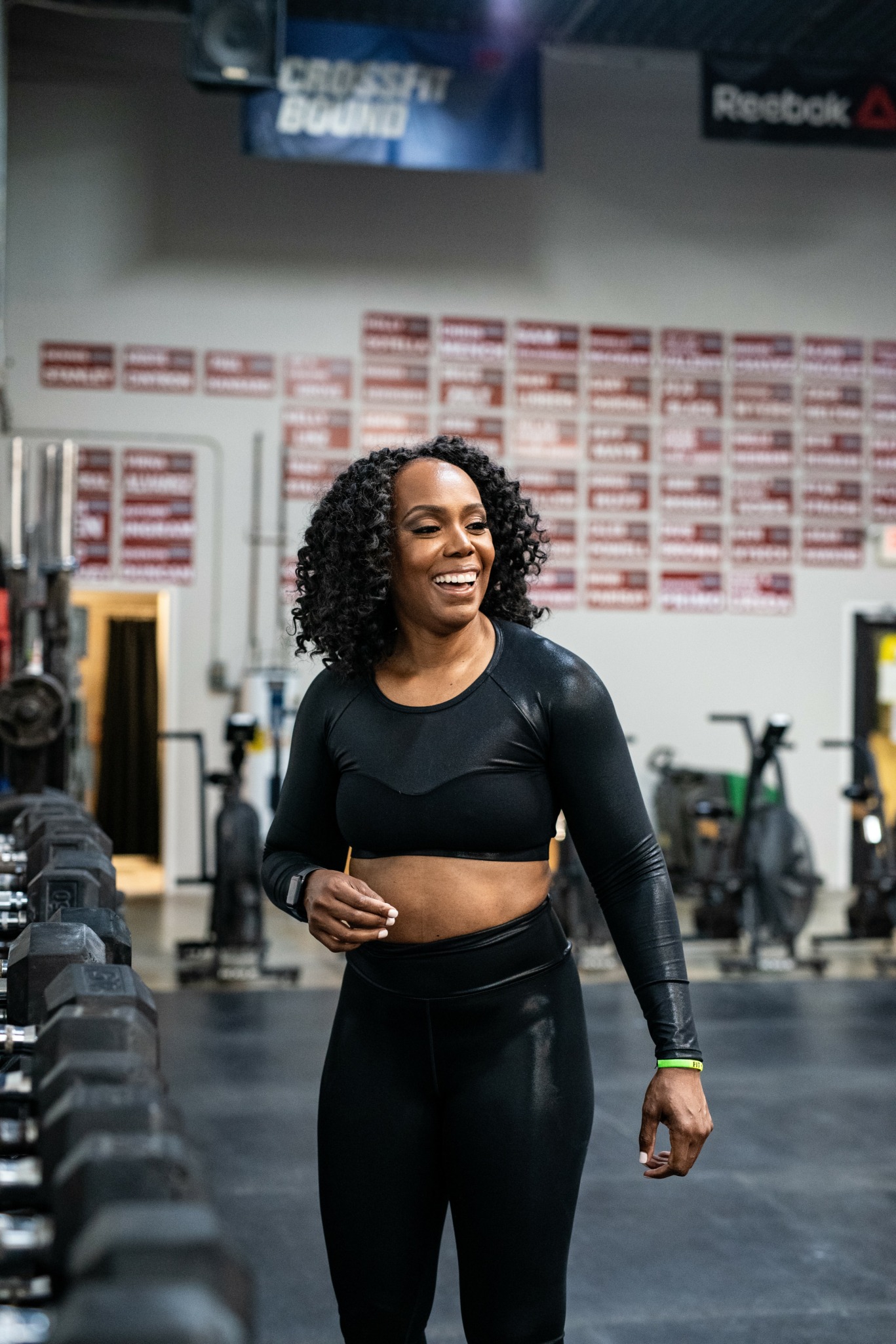 Smiling woman with curly hair in workout clothes standing in gym with equipment and posters.