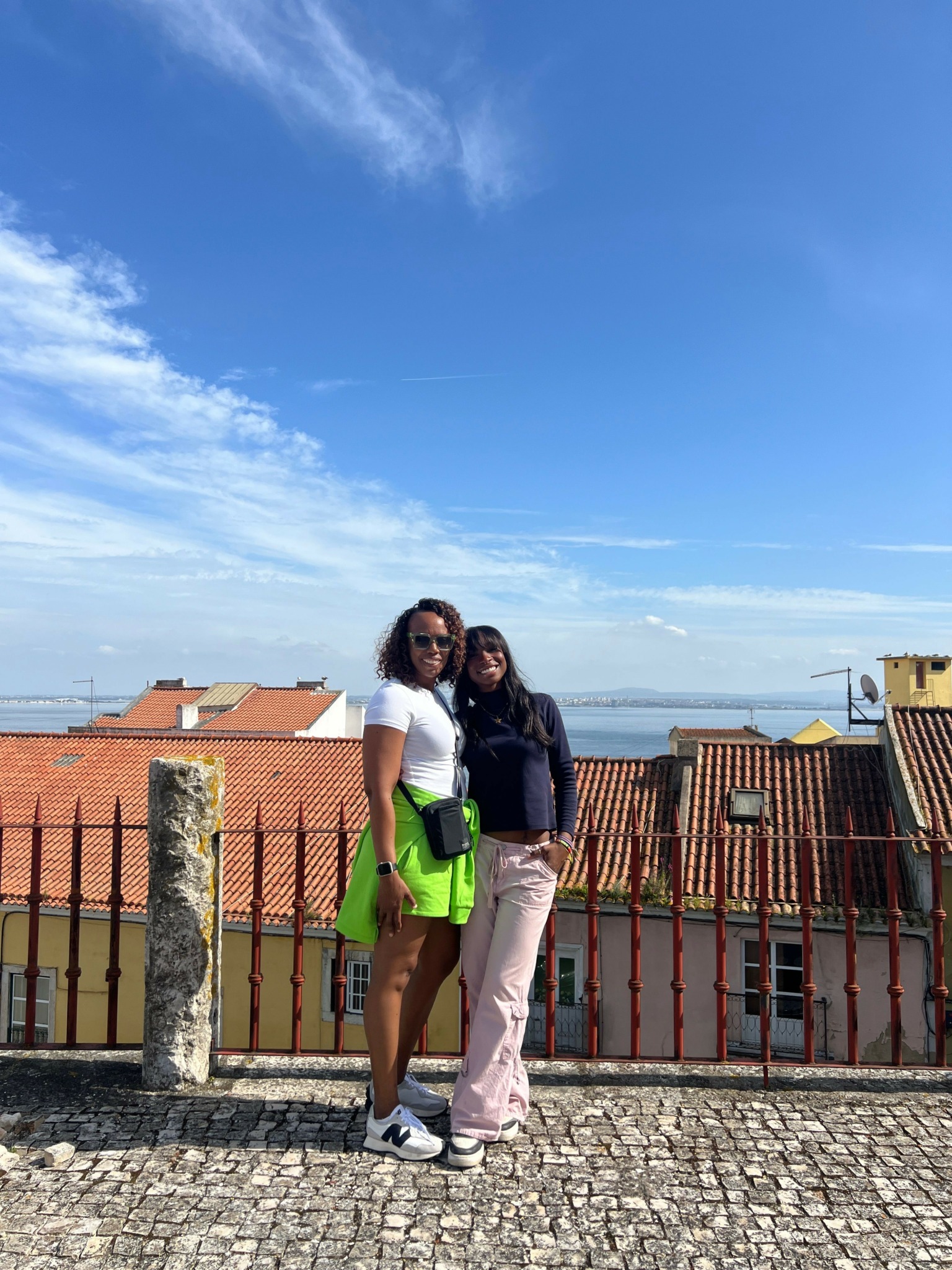 Two women standing on cobblestone street near a fence and buildings under a blue sky with clouds.
