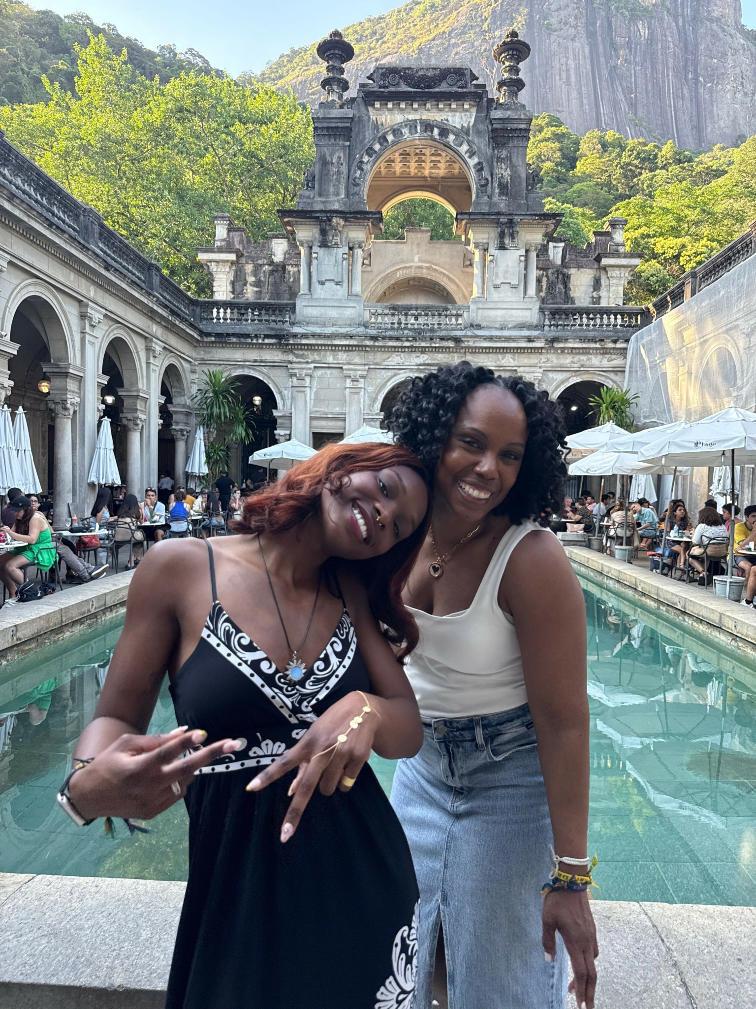 Two women smiling and posing together in front of a fountain and historic building, with trees and people in background.