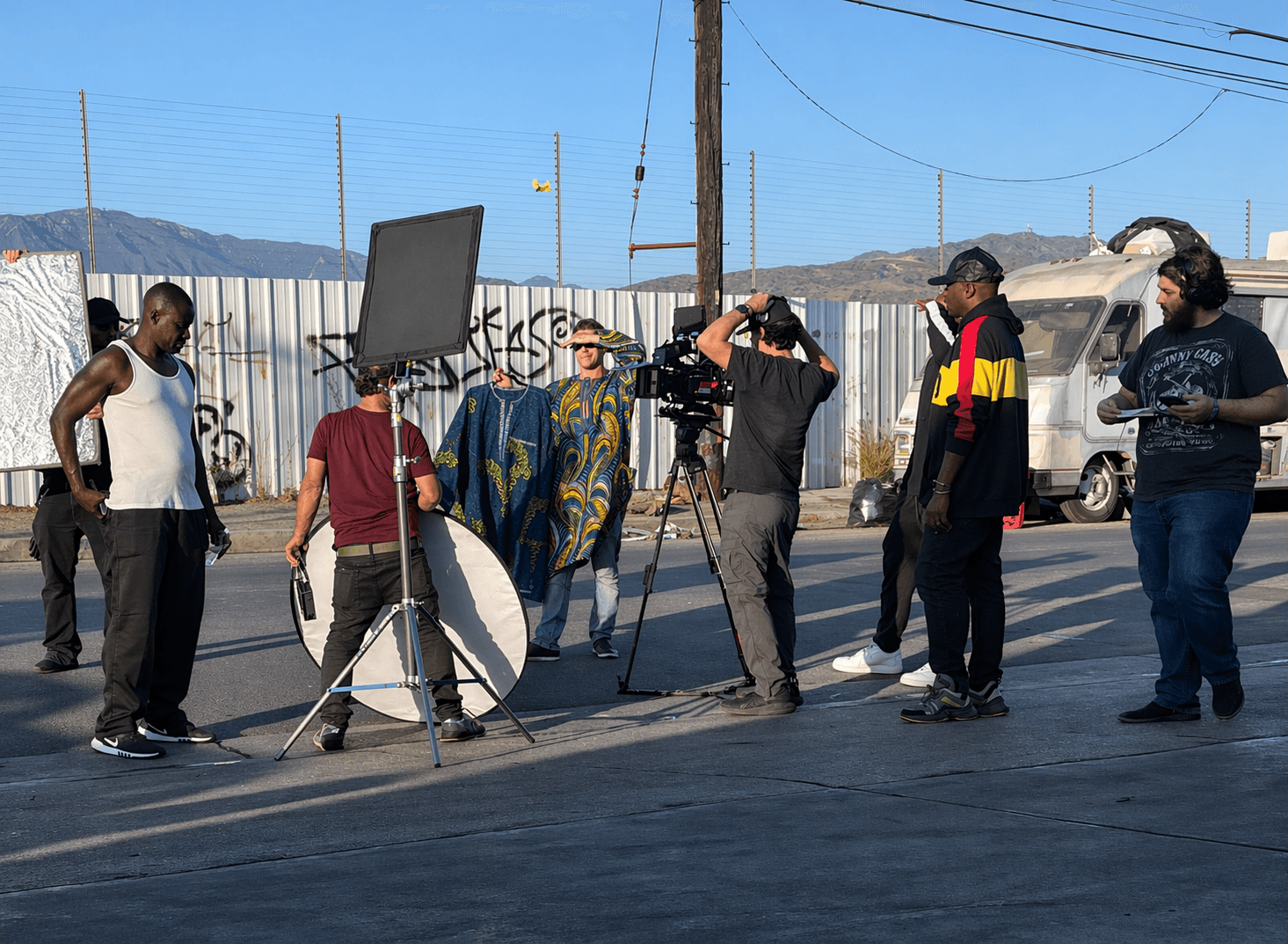 Group of people filming a video outdoors with equipment, in a sunny area with a fence and vehicles nearby.