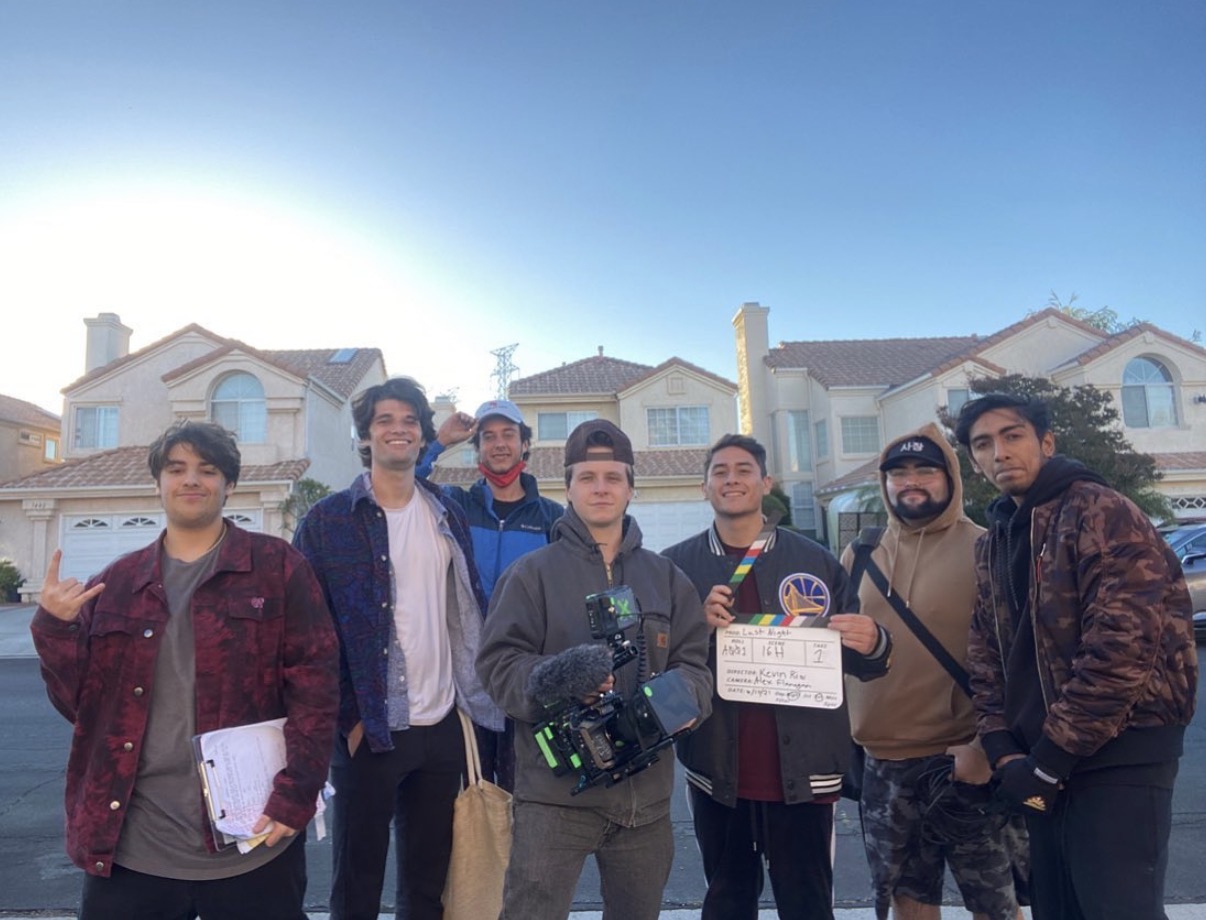 Group of seven people standing outdoors in front of houses, some holding items, with a bright sky behind.