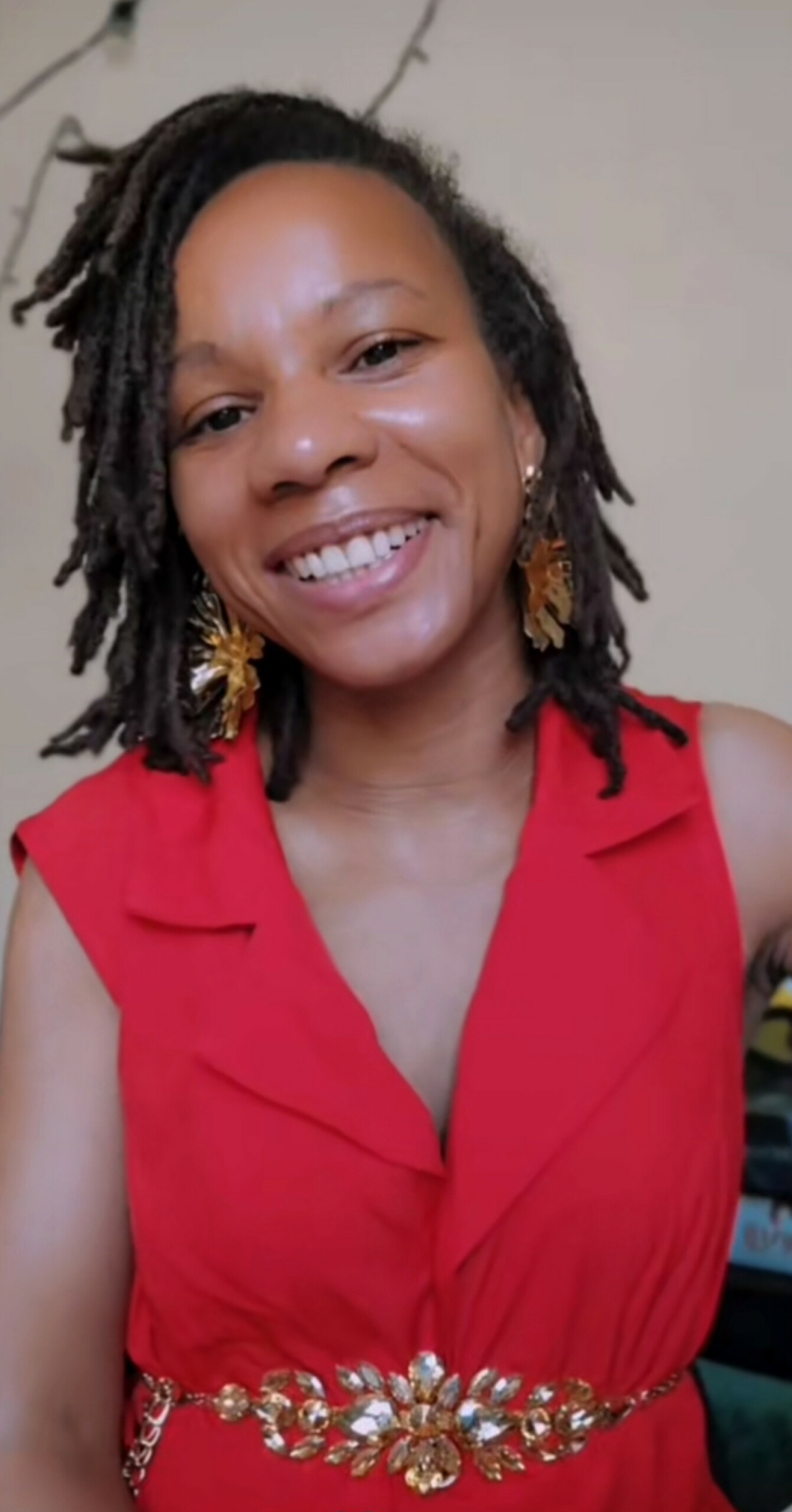 Smiling woman with dreadlocks wearing a red dress and gold earrings.