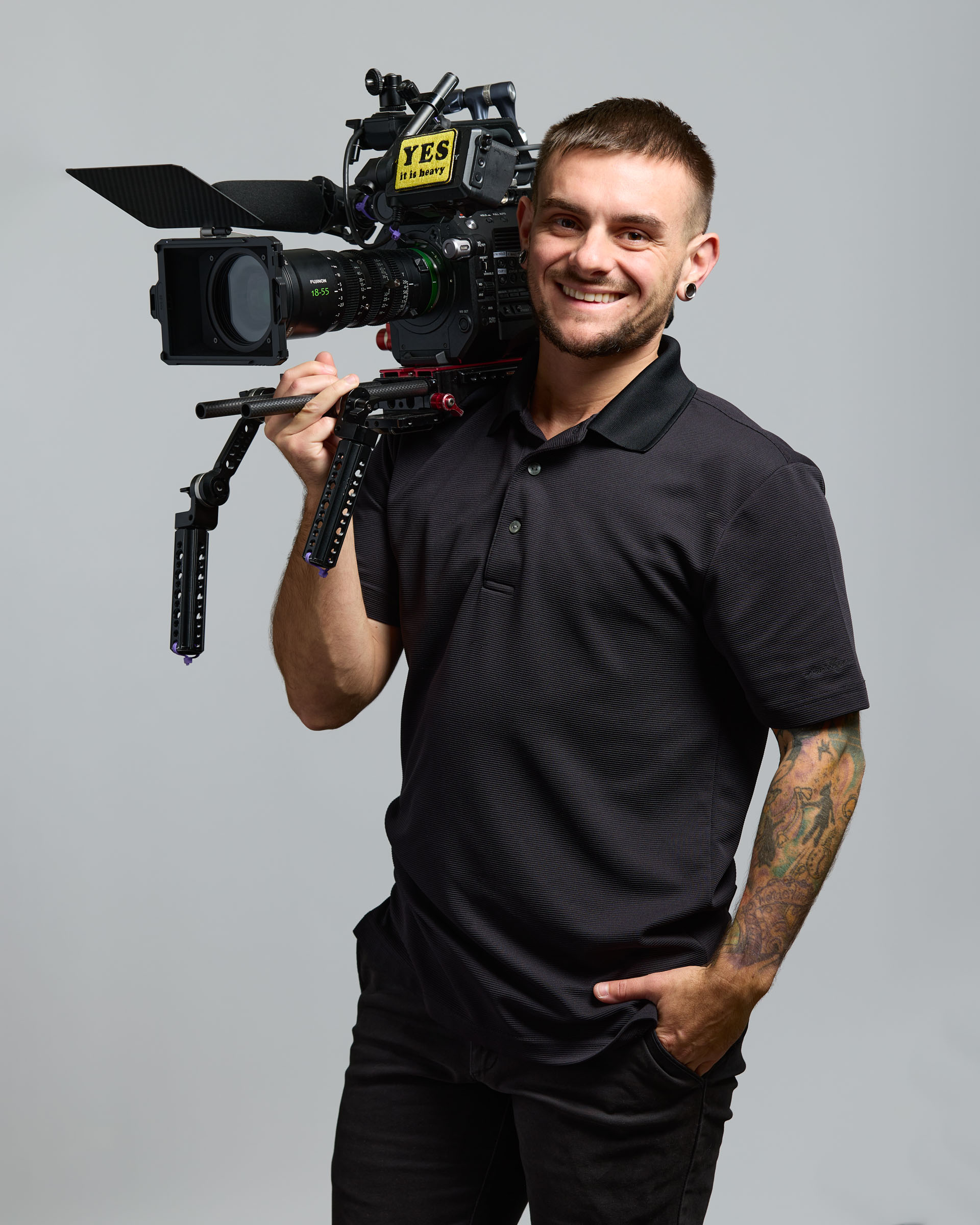 Young man with short hair and tattoos, smiling, holding a professional video camera on his shoulder, wearing a black polo shirt.