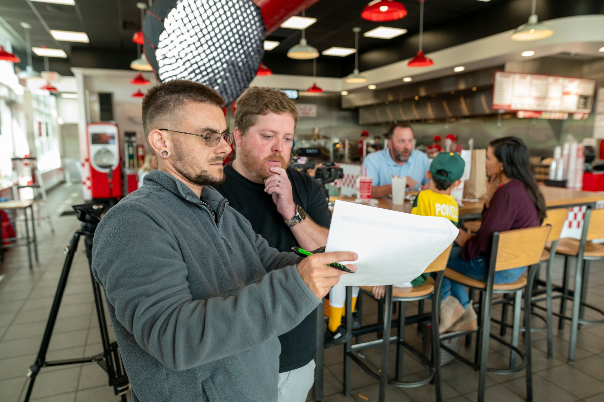 Two men review a large sheet of paper in a restaurant or cafe setting with people sitting at tables in the background.