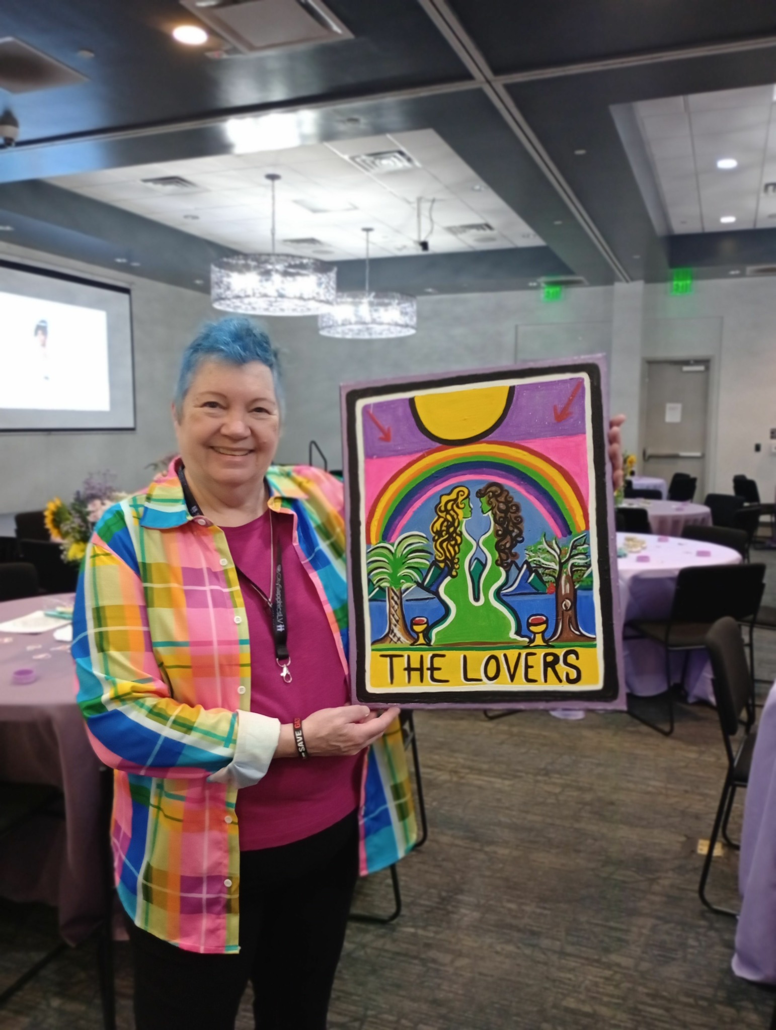 Person with colorful striped jacket holding a sign with a rainbow and cactus design, reading 'The Lovers'.