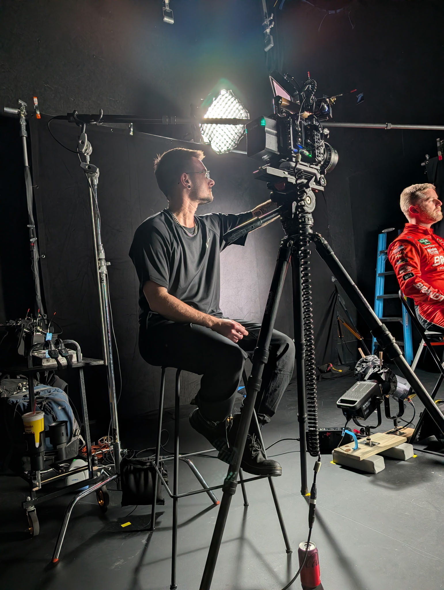 Person sitting on a stool operating a camera on a tripod in a studio setting with lighting equipment.