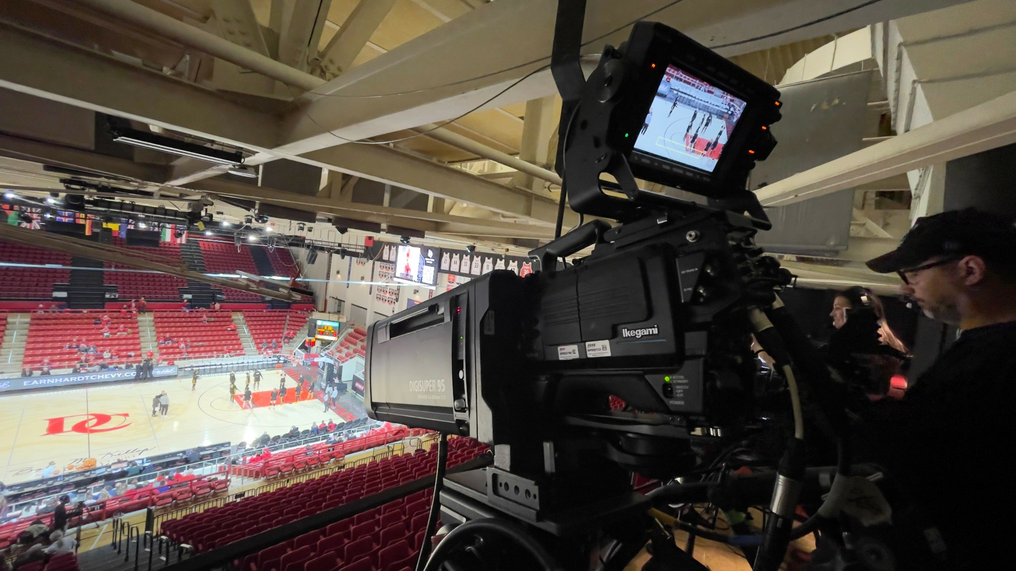 Camera operator filming an indoor sports arena with red seats and a large screen.