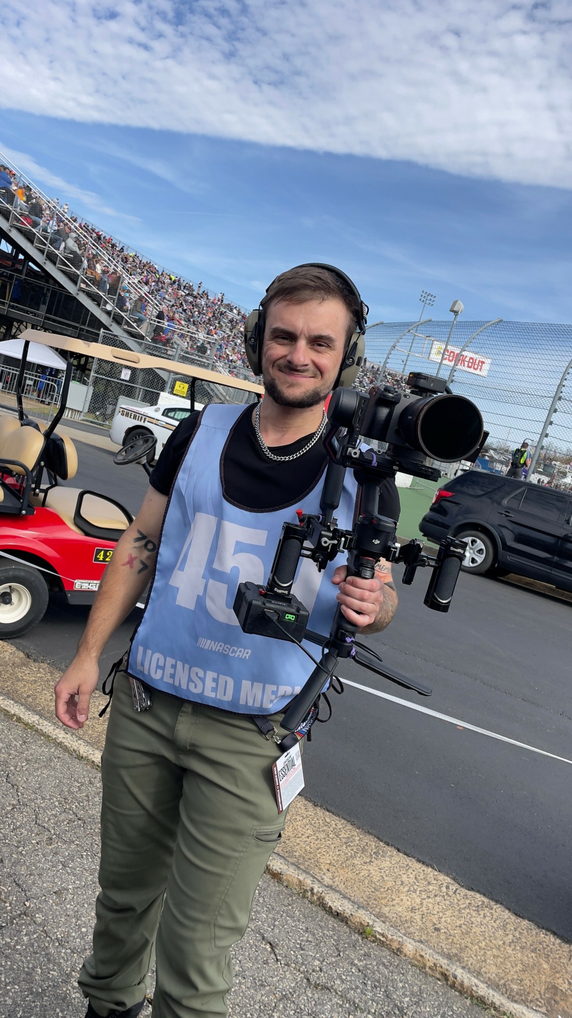 Man with headphones holding a camera on a tripod, standing outdoors near a race track with cars and spectators.