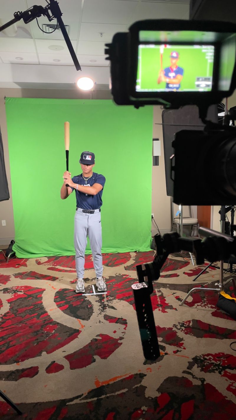 Person in baseball uniform holding a bat in front of a green screen, camera capturing the scene, patterned carpet below.