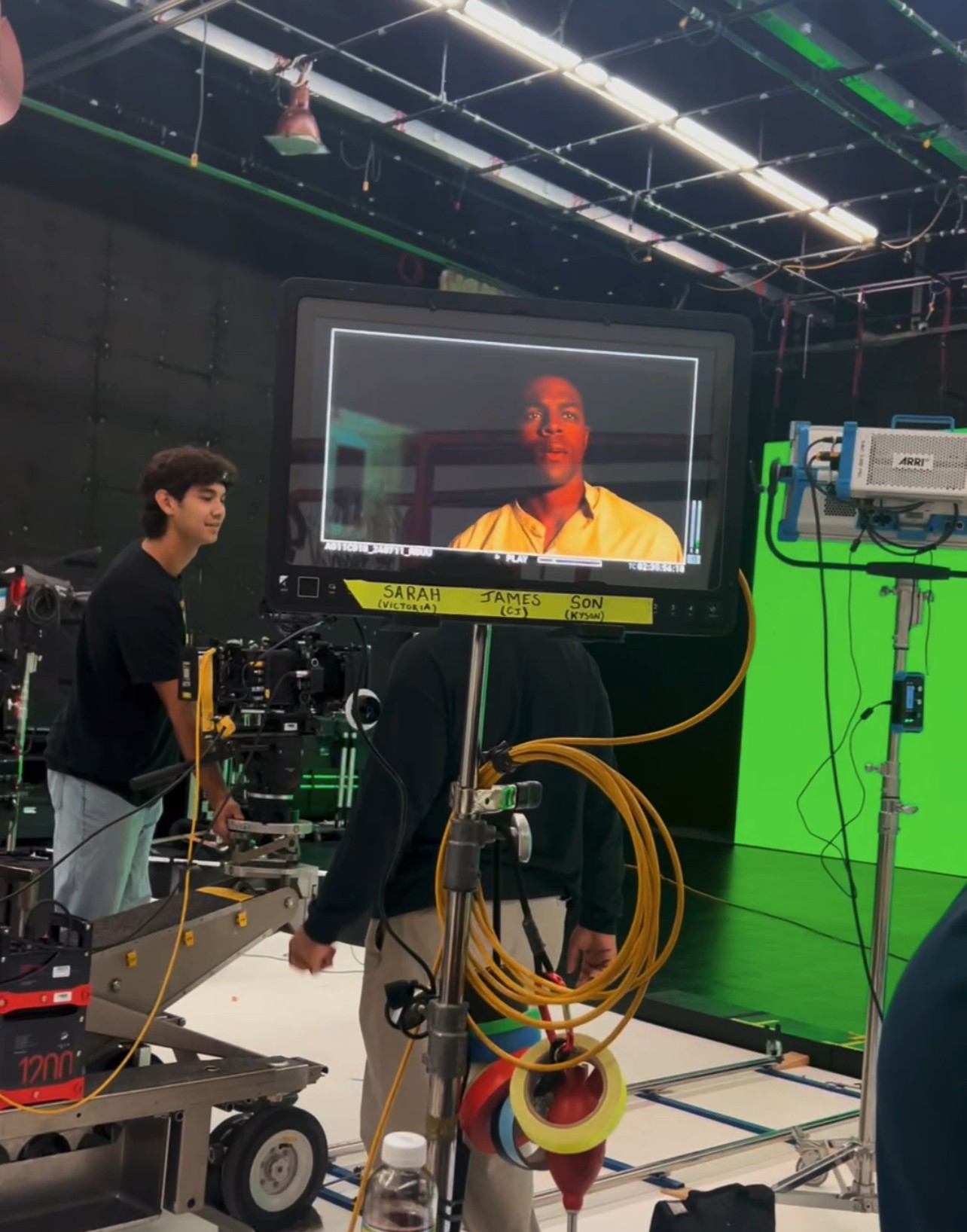 Person operating camera equipment in a studio with a large monitor displaying a person's face, green screen, and studio lights.