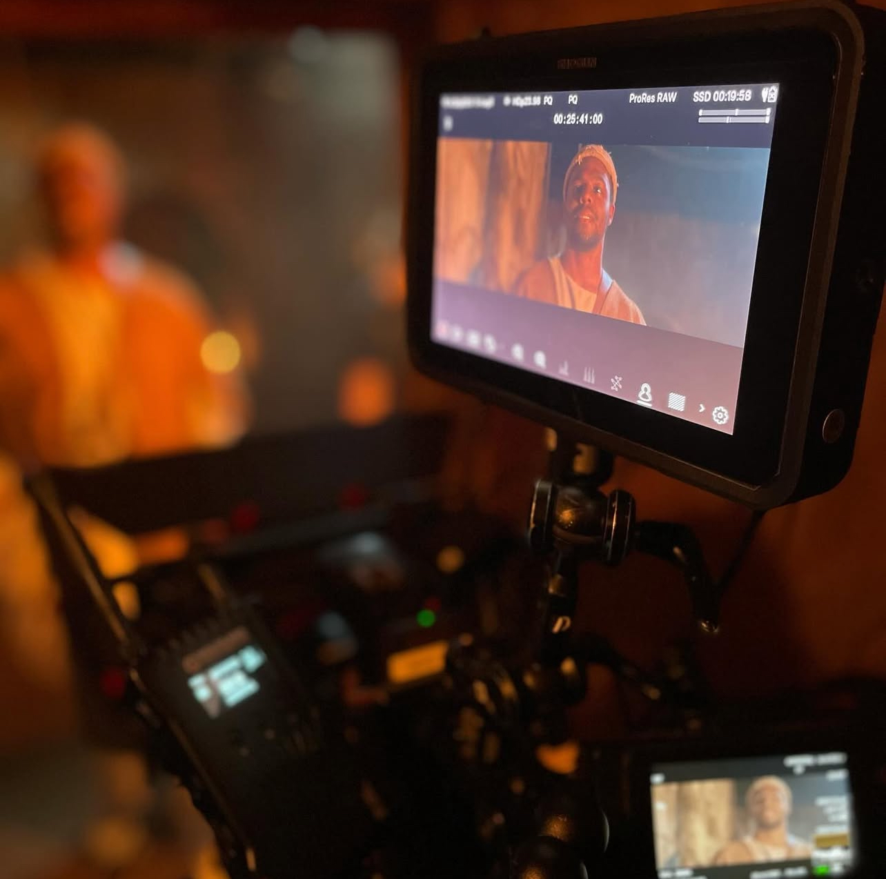 Camera monitor shows a person with a head covering, on set with blurred background and warm lighting.