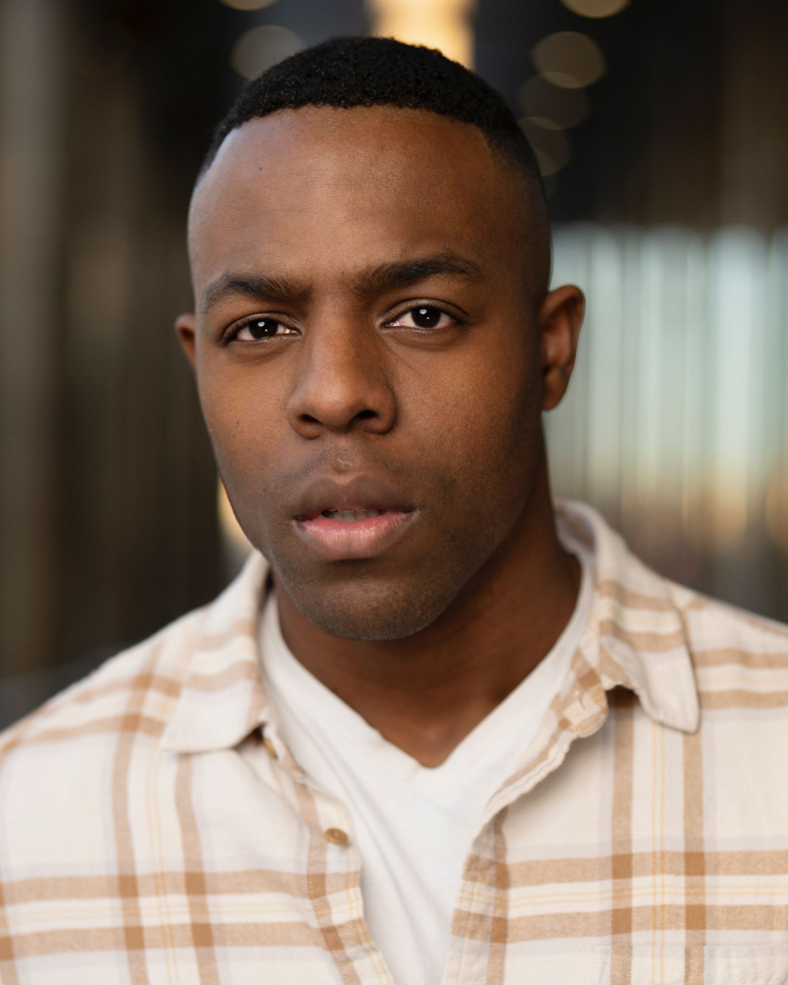 Close-up of a man with short hair, wearing a plaid shirt and white t-shirt, looking directly at the camera.