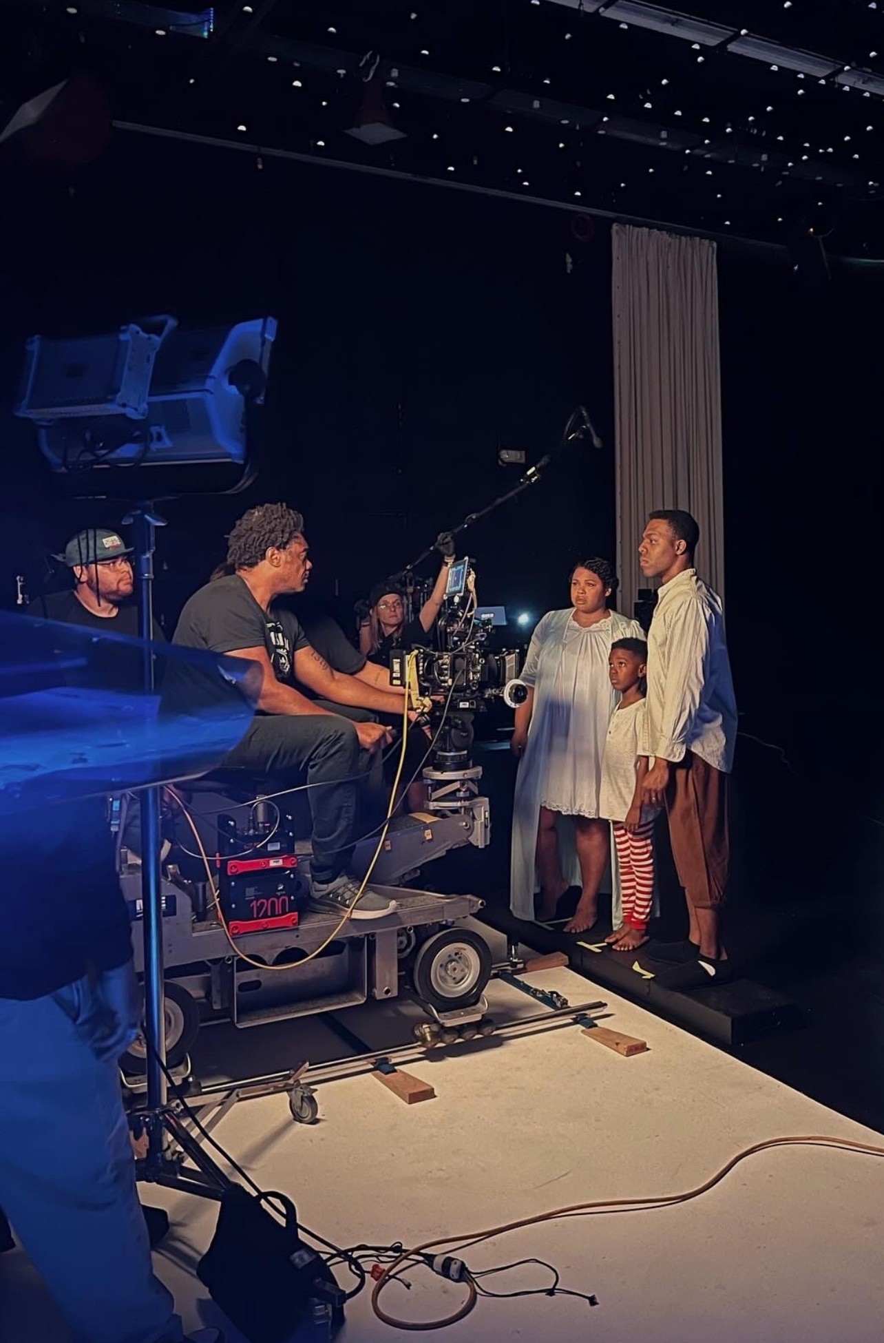 People on a stage with camera equipment, some children and adults, in a dark room with star-like ceiling lights.
