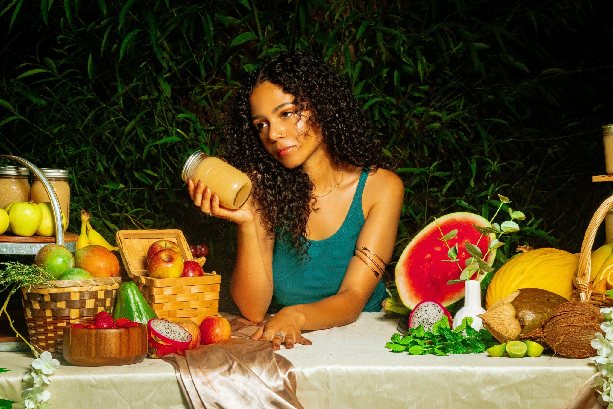 Woman with curly hair sitting at a table with fruits and a watermelon, holding a jar, outdoors at night.
