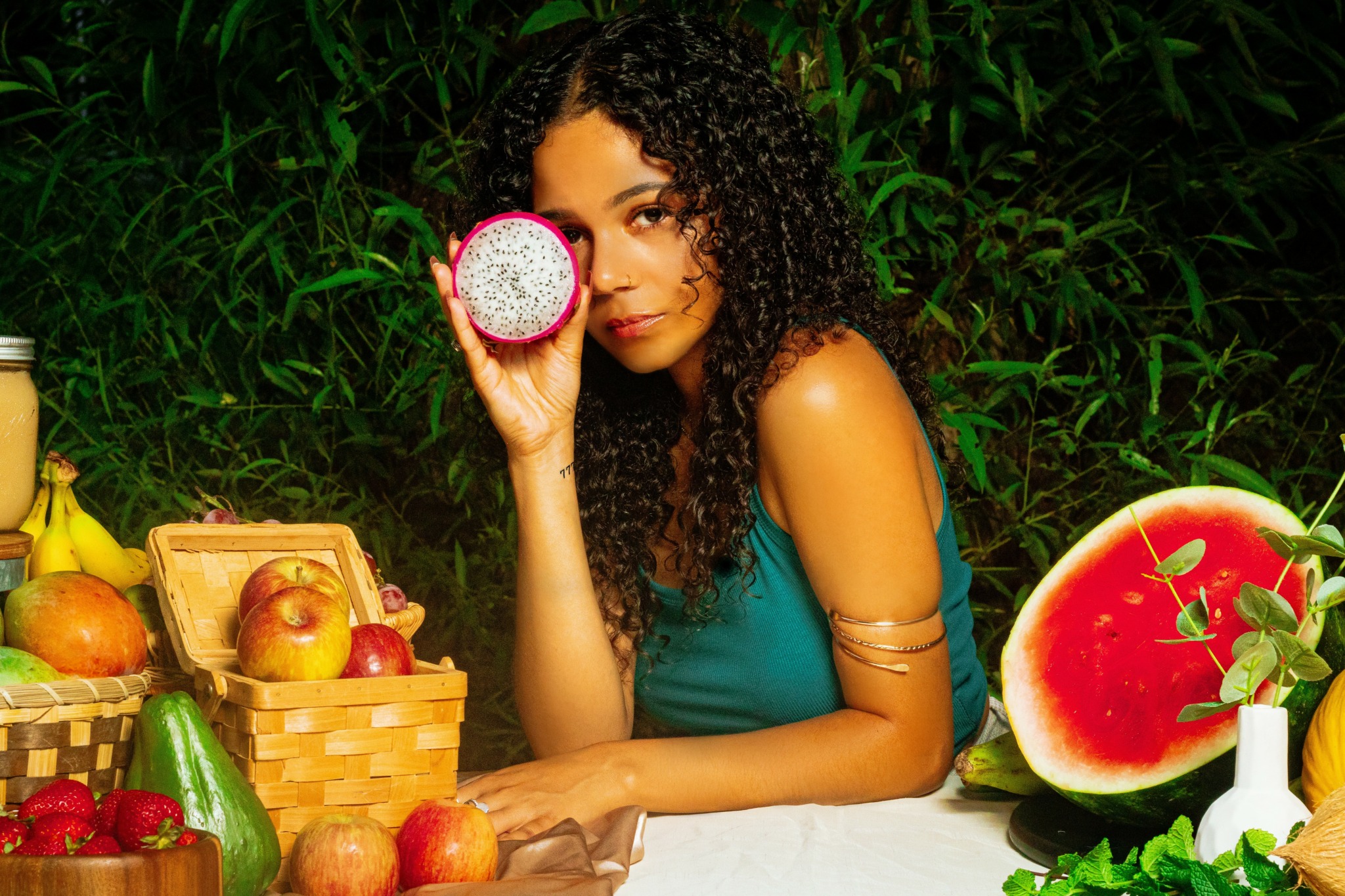 Woman with curly hair holding a pink and white donut near her eye, sitting at a table with fruits and a watermelon.