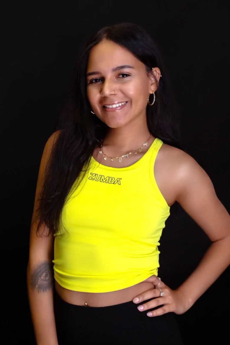 Young woman with long dark hair smiling, wearing a bright yellow sleeveless top and black pants, standing against black background.