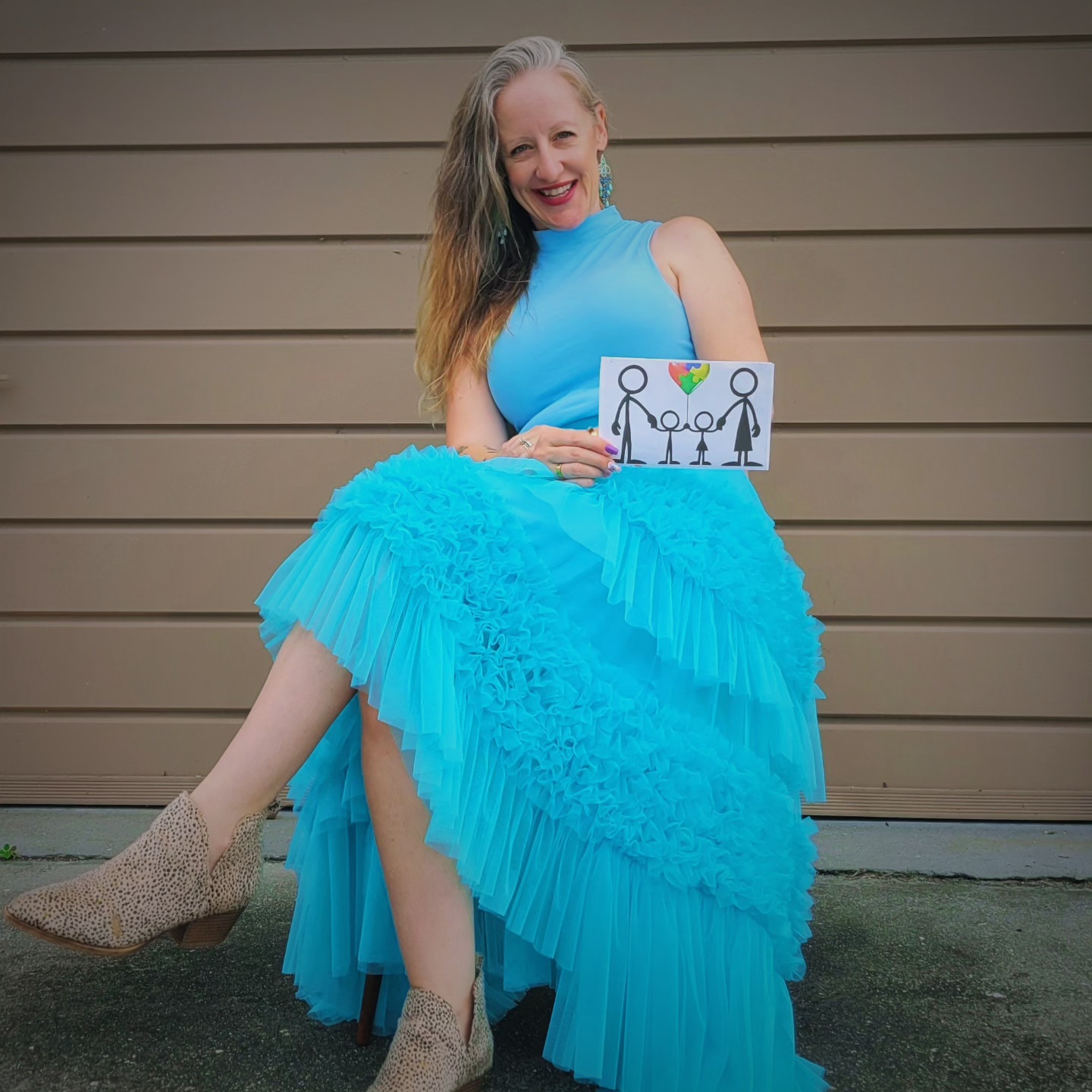 Woman in a blue dress sitting on a chair, holding a drawing of people holding hands, smiling.