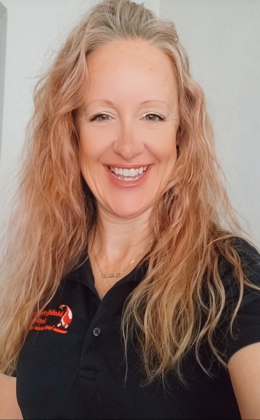 Woman with long, wavy blonde hair smiling, wearing a black polo shirt with a logo.