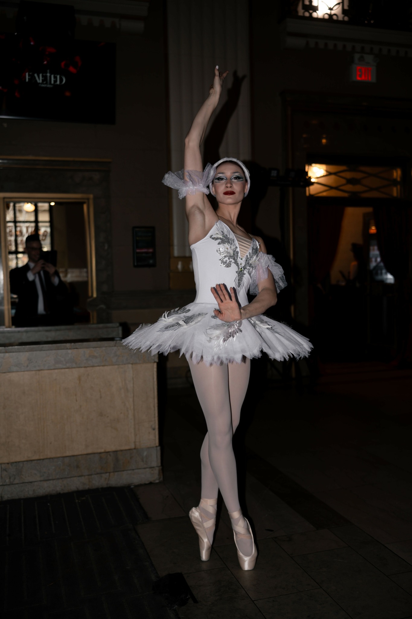 Ballet dancer in tutu and pointe shoes poses with one arm raised in a dimly lit room.