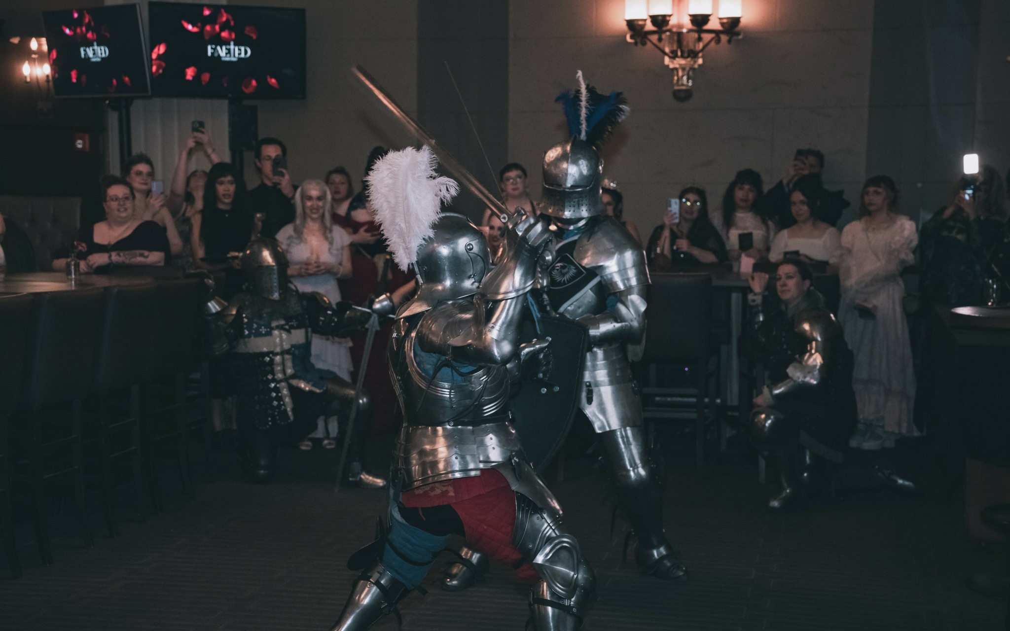 Two people in knight costumes fighting in a dimly lit room with an audience watching.