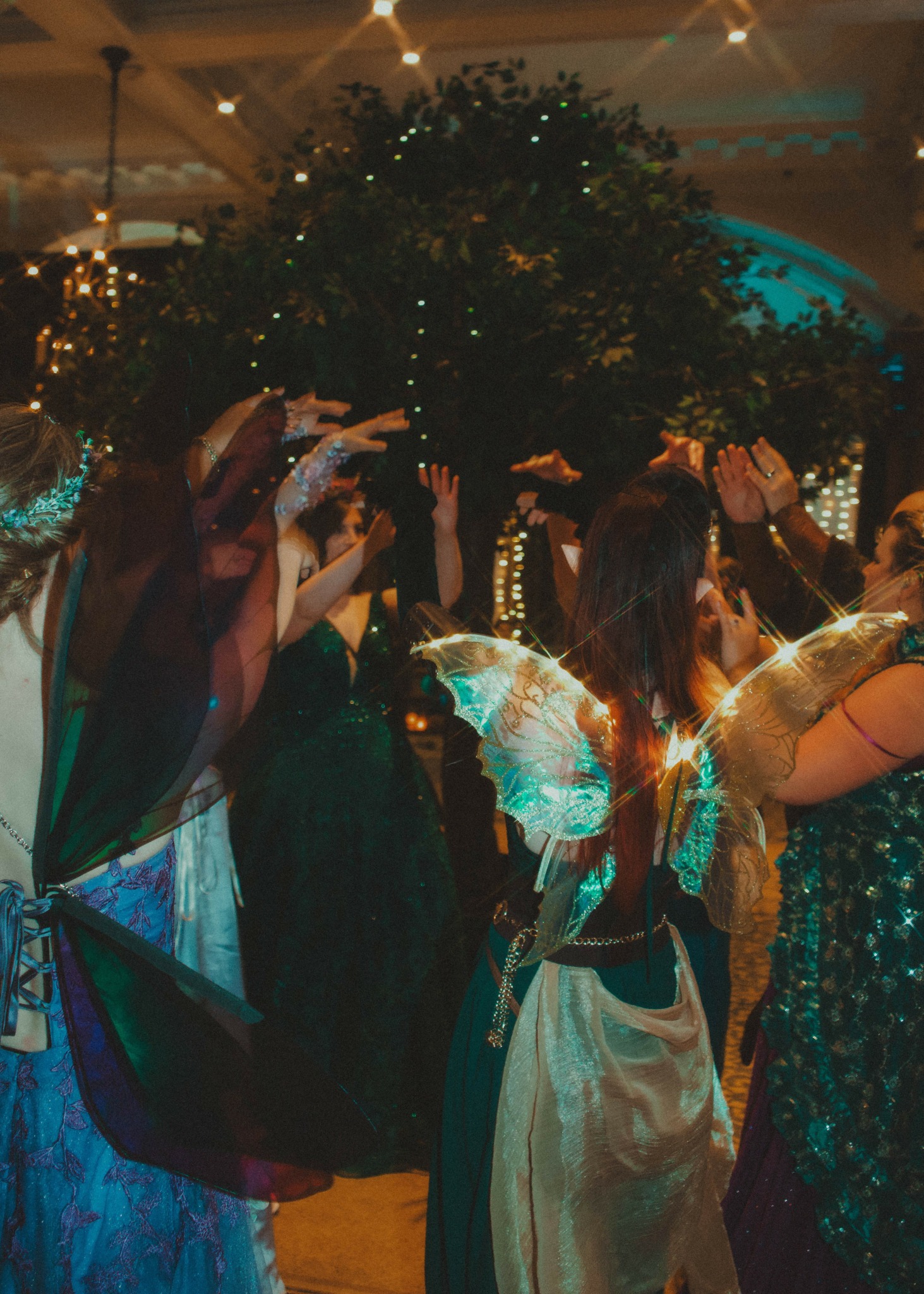 Group of people dancing indoors with a large decorated tree in the background, some with hands raised.