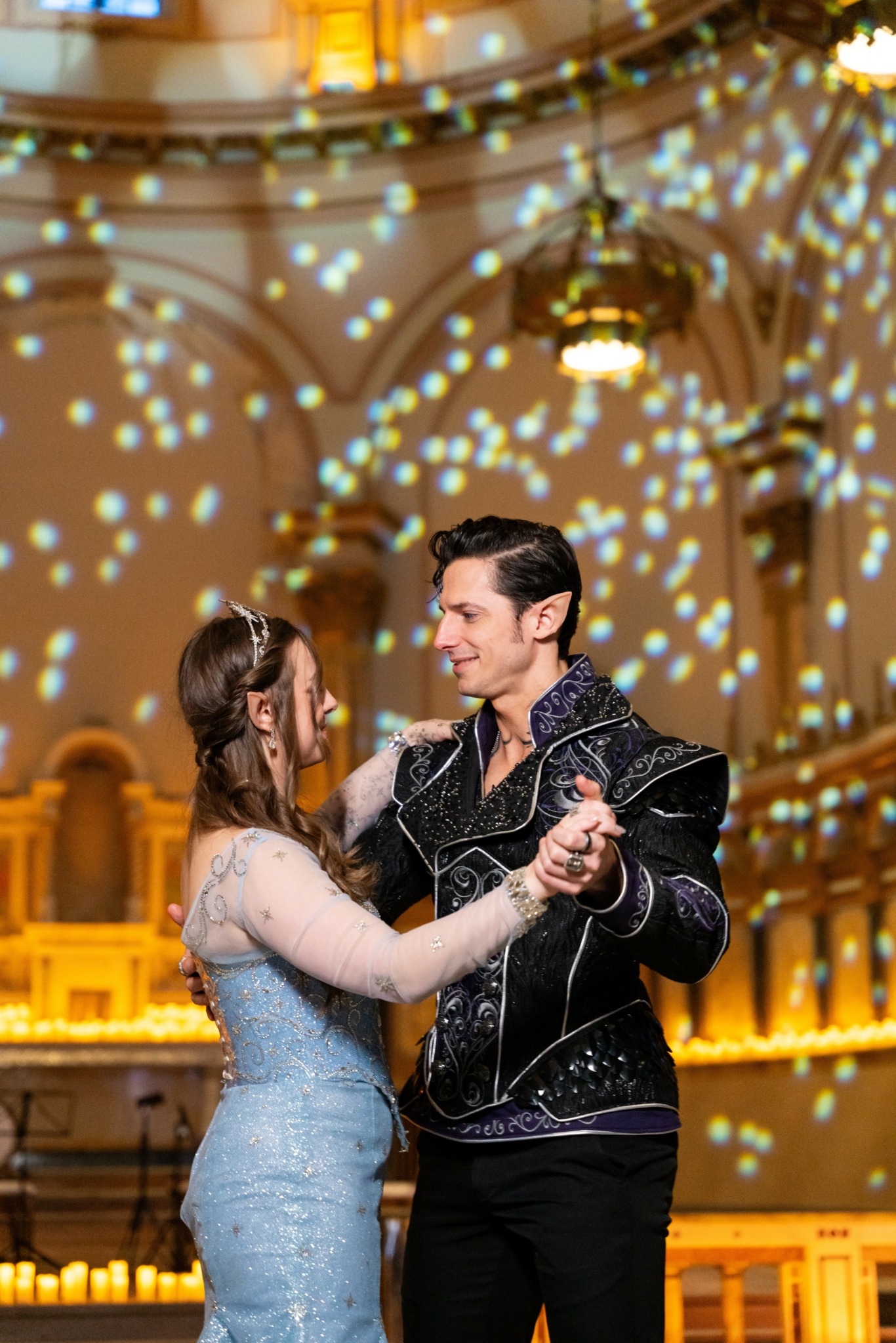 A couple dancing in a grand hall with ornate architecture and hanging lights, surrounded by sparkling light effects.