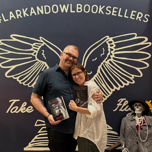 Two people smiling and holding books in front of a dark background with a large winged design and text.