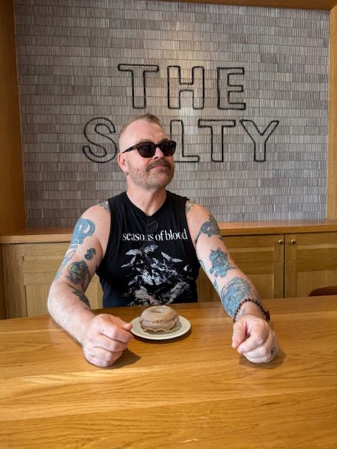 Man with tattoos and sunglasses sitting at a wooden table with a bowl, in front of a wall with 'THE SKY' sign.