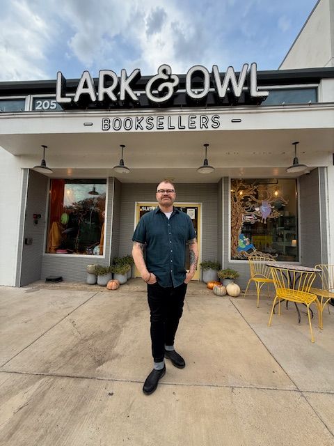 Man standing outside bookstore with yellow chairs and pumpkins, storefront with large window, sidewalk, cloudy sky.