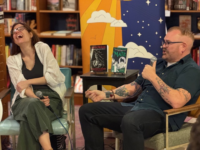 Two people sitting and laughing in a room with bookshelves and colorful backdrop, one holding a microphone.
