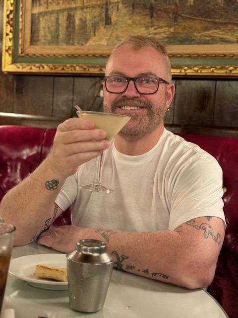 Man with glasses and tattoos holding a cocktail glass, smiling, sitting at a table in a restaurant.