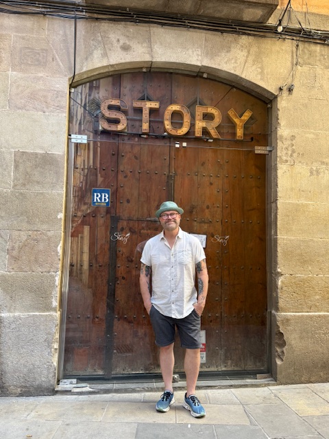 Person standing in front of a large wooden door with the word 'STORY' above it, stone archway, casual clothing, glasses, cap.