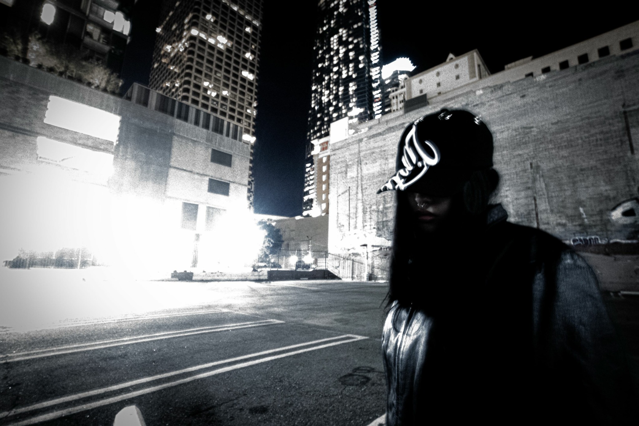 Person wearing a helmet and jacket standing on city street at night with tall buildings in background.